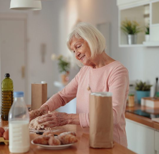 Happy senior woman housewife making kneading dough for biscuits while standing behind breakfast bar in kitchen